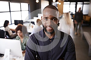 Bearded black male creative standing in a busy casual office, smiling to camera