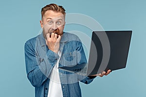 Bearded blonde hipster young man holding laptop with scary face over blue background.