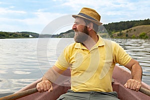 A bearded boatman in a hat looks at the shore while sitting in a boat and holding the oars in his hands