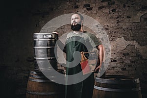 Bearded brewer in apron holds the wooden box with beer while standing near barrels at brewery factory.