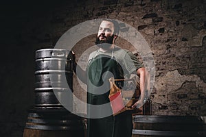 Bearded brewer in apron holds the wooden box with beer while standing near barrels at brewery factory.