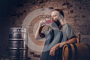 Brewer in apron standing near barrels and drinks craft beer at old brewery factory.