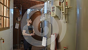 Bearded brewer man on a background of beer tanks and a brewery making notes in a tablet