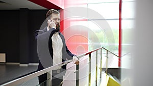 Bearded brunette businessman is going up the stairs and coming to glass fence while talking on the phone inside modern