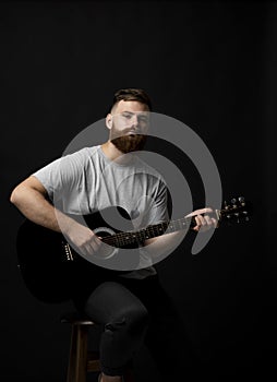 Bearded brutal guitarist plays an acoustic guitar in a black room and looking in the camera. The concept of music