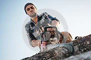 Bearded brutal lumberjack wearing plaid shirt sawing tree with chainsaw for work on sawmill