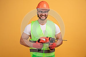 Bearded builder in helmet, reflective vest, glasses and gloves holding a drill in his hands.  on yellow background. Yes, I