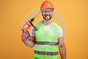 Bearded builder in helmet, reflective vest, glasses and gloves holding a drill near his head. fools at work. Isolated on yellow