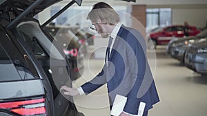 Bearded businessman choosing the car in motor show close up. Tall man holding tablet looks in opened trank and examines