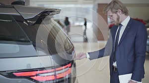 Bearded businessman choosing the car in motor show close up. Tall man holding tablet opens trunk and examines vehicle