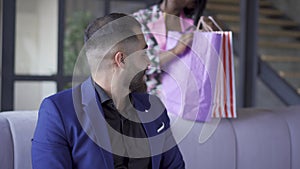 Bearded businessman sitting on the sofa at home looking at camera. Happy African American woman back home from shop with