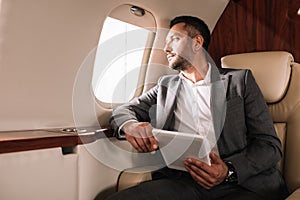 bearded businessman in suit holding digital tablet while looking at airplane window in private jet.