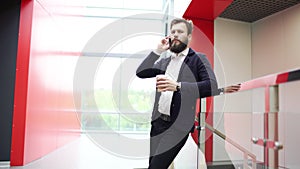 Bearded businessman is talking on the phone and holding cup of take-out coffee while standing inside modern urban office