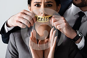 Bearded businessman touching scotch tape with sexism lettering on mouth of scared businesswoman gesturing