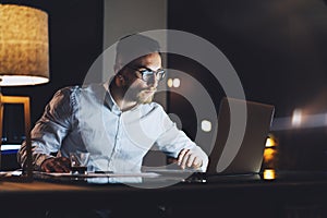 Bearded businessman wearing white shirt working on modern loft office at night. Man using contemporary notebook texting