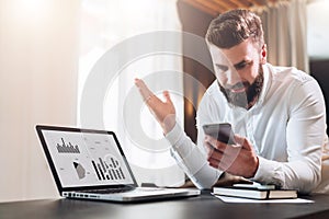 Bearded businessman in white shirt is sitting at table in front of laptop with graphs, charts, diagrams on screen