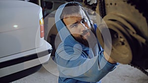 The bearded car mechanic sits and from below screws the bolt into the body of the motor hanging on the chains.