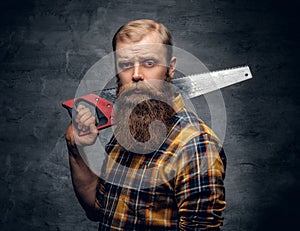 Bearded carpenter dressed in a plaid shirt holds handsaw.