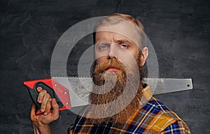 Bearded carpenter dressed in a plaid shirt holds handsaw.