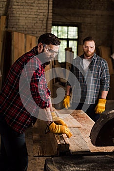 bearded carpenter in safety googles using machine saw and partner standing behind