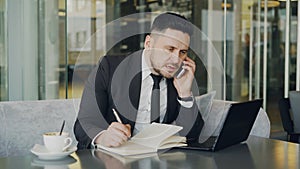 Bearded Caucasian businessman using laptopc omputer, talking on smartphone and writing down business information in cafe