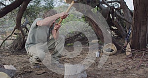 Bearded caucasian male survivalist using axe to cut fire logs at camp in wilderness