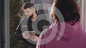 Bearded caucasian police officer asking questions and looking around. Unrecognisable brunette woman standing at entrance