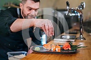 Bearded chef cook dressed black uniform sprinkling the sushi Nigirizushi dish in basement restaurant kitchen. Professional