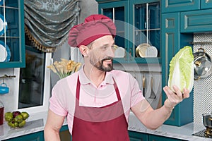 Bearded Chef in red apron and hat holds and looks at napa cabbage on kitchen. pakchoi in hand of cook. Fresh Chinese leaves tested