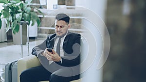 Bearded concentrated businessman using smartphone sit on armchair inside luxury hotel after arrivel to business meeting