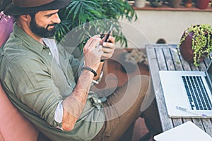 Bearded confident hipster wearing green shirt and brown hat texting message via smartphone on terrace outside. Positive