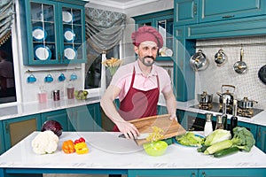 Bearded cook cutting yellow paprika. Smiling guy in cap makes salad with paprika.