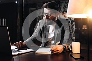 Bearded coworker sits by the wooden table with lamp and using contemporary notebook. Businessman hold pen and analysing trends