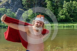 Bearded elderly man in a Santa Claus hat with a red towel. Against the background of a summer landscape
