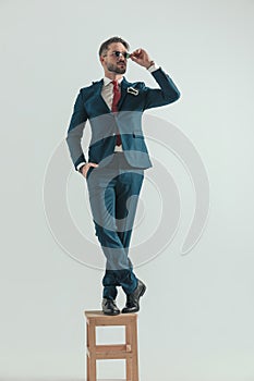 Bearded elegant man in suit standing on wooden chair and adjusting glasses