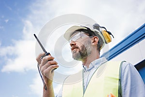 Bearded engineer wearing helmet using walkie-talkie to communicate with industry produce site