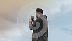 Bearded European man stands with smartphone. Low angle amazing sky view. Casual handsome male takes smartphone photo. 4K
