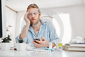 Bearded fair-haired male office worker looking unhappily at screen of smartphone, leaning on his elbow, sitting at table