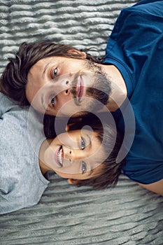bearded father and his son are lying on the bed with phone and taking selfies at the window in a white room