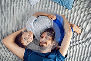 bearded father and his son are lying on the bed with phone and taking selfies at the window in a white room