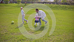 Bearded father plays at the stadium in football with two daughters. Summer day, green grass.