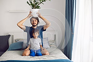 bearded father and son in T-shirts are sitting on a bed. man is holding a pot with flower on his head in a white room by