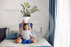 bearded father and son in T-shirts are sitting on a bed. man is holding a pot with flower on his head in a white room by