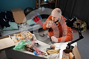 Bearded freelancer man working at home, eating pizza in Messy, cluttered room with piles garbage