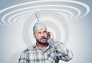 Bearded funny man in an aluminum hat talking on a smartphone with the sky, taking cosmic electric waves, concept idiots