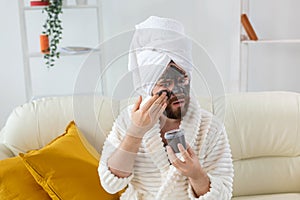 Bearded funny man having fun with a cosmetic mask on his face made from black clay. Men skin care, humor and spa at home