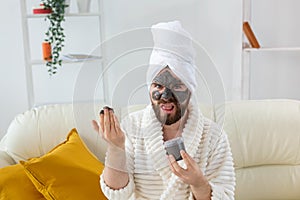 Bearded funny man having fun with a cosmetic mask on his face made from black clay. Men skin care, humor and spa at home