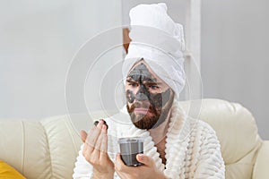 Bearded funny man having fun with a cosmetic mask on his face made from black clay. Men skin care, humor and spa at home