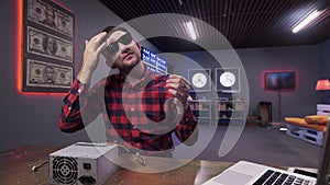 Bearded guy touches his hair having box of power supply with cooler put on desk.