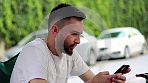 Bearded and handsome young man using mobile phone outdoors. Serious man in white t-shirt looking at mobile phone screen and readin